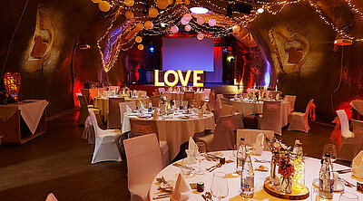 Die Event-Kaverne ist fertig eingerichtet und bereit für die Feier. Auf der Bühne steht ein grosser "LOVE" Schriftzug welcher mit Lampen beleuchtet ist.